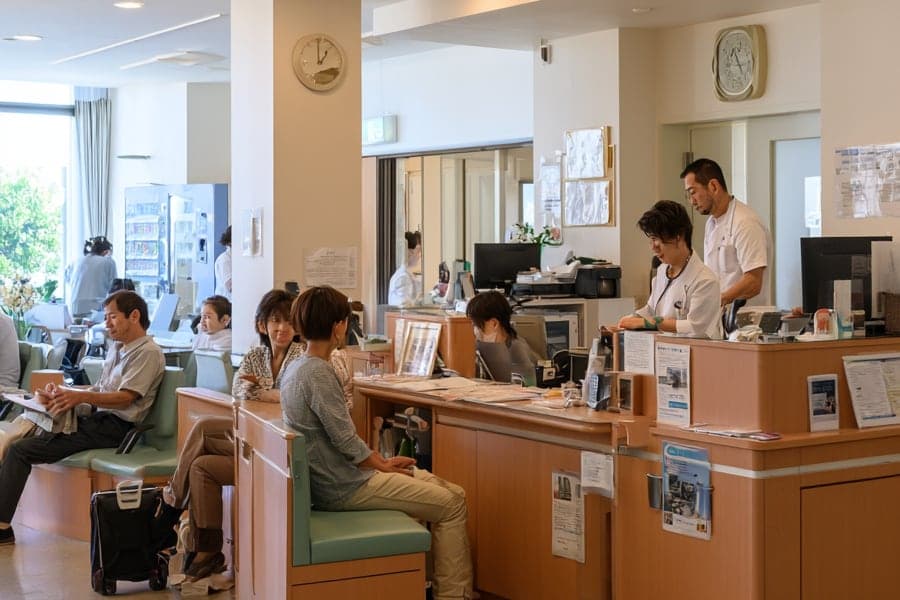 歯科医院の受付と待合。木のカウンターの内側に白衣のスタッフが並び、左手の淡い緑の長椅子で数人が順番を待っている。壁に掛け時計。